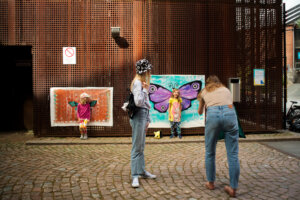 Kaksi lasta seisoo taideteoksien edessä, etualalla aikuinen ottaa heistä kuvan.
